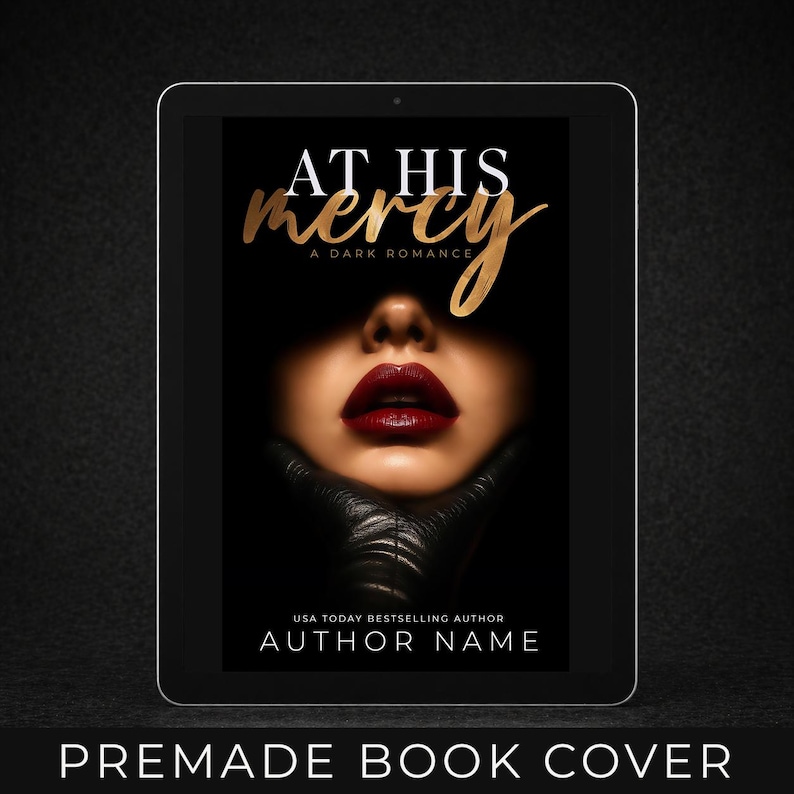 May include: A digital tablet displays a premade book cover with the title "At His Mercy" in gold script. The cover features a woman's face with dark red lipstick and a black leather glove. The text "A Dark Romance" and "Author Name" are also visible.