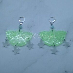 May include: A pair of green butterfly earrings with silver star charms hanging from them. The earrings are made of resin and have a translucent finish.