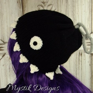 May include: Black knitted beanie hat with a white eye and white teeth around the brim. The hat has a grey drawstring.