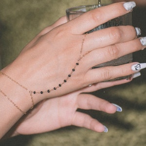 May include: A delicate gold hand chain with small black beads. The chain connects to a ring and wraps around the hand. The nails are painted with a white and gray design, and one nail has a black and white graphic.