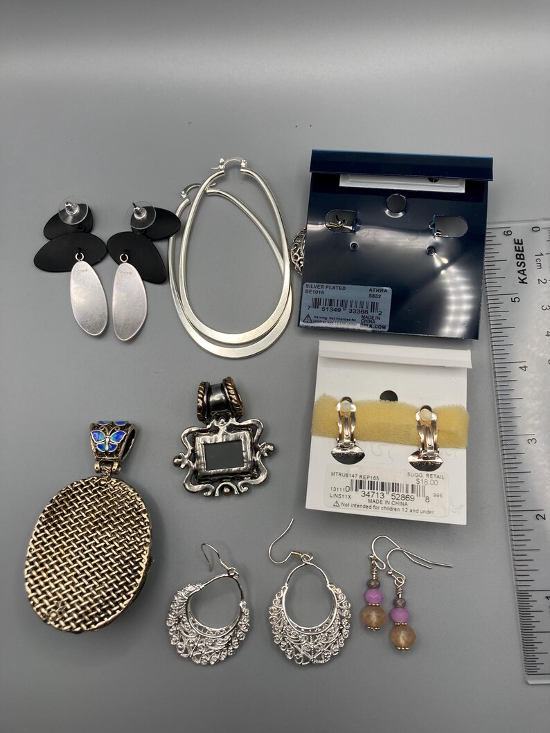 May include: A collection of jewelry including silver-toned hoop earrings, black and silver drop earrings, two pendants, and two pairs of earrings on display cards. A ruler is visible on the right, showing a scale of approximately 5.5 inches in length.