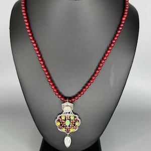 May include: A red beaded necklace with a silver pendant. The pendant is adorned with colorful gemstones in shades of red, green, and yellow. A teardrop-shaped crystal hangs from the bottom of the pendant. The necklace is displayed on a black mannequin.
