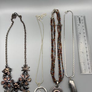 May include: Four necklaces arranged vertically next to a 6-inch ruler. From left: a dark metal floral bib necklace, a silver-tone chain with a blue cabochon circle pendant, a multi-strand brown beaded necklace with an oval turquoise-style pendant, and a silver-tone chain with a long, smooth, metallic pendant.