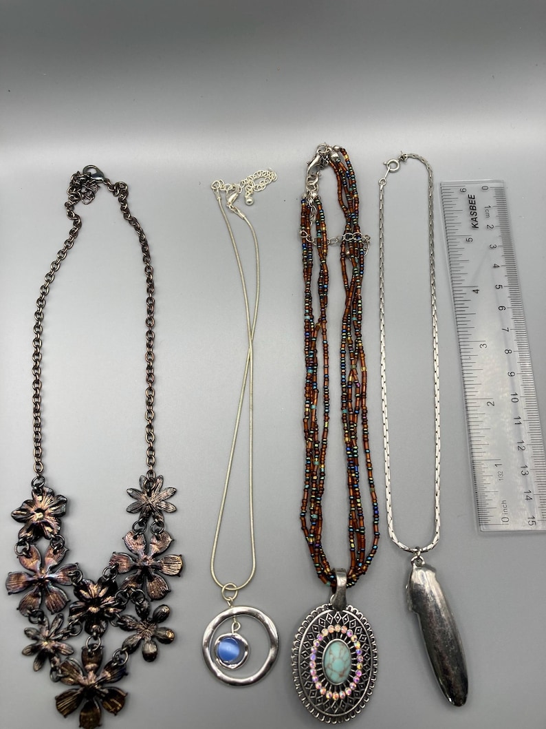 May include: Four necklaces arranged vertically next to a 6-inch ruler. From left: a dark metal floral bib necklace, a silver-tone chain with a blue cabochon circle pendant, a multi-strand brown beaded necklace with an oval turquoise-style pendant, and a silver-tone chain with a long, smooth, metallic pendant.