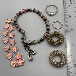 May include: A collection of jewelry including a pink enamel flower bracelet, a multi-colored beaded bracelet with metallic spacers, a silver band ring with a small stone, a twisted silver ring, and two silver-toned spiky sensory rings next to a ruler.
