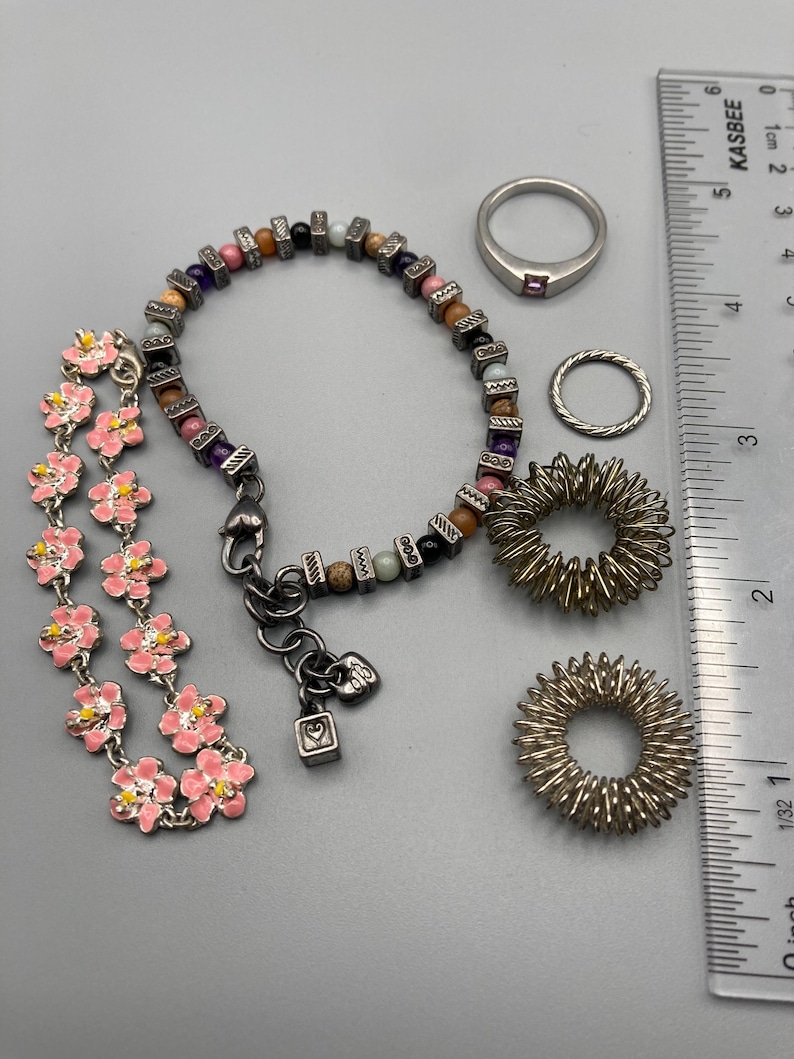May include: A collection of jewelry including a pink enamel flower bracelet, a multi-colored beaded bracelet with metallic spacers, a silver band ring with a small stone, a twisted silver ring, and two silver-toned spiky sensory rings next to a ruler.