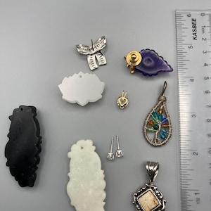 May include: A collection of jewelry pieces including a silver bow earring, a purple leaf-shaped pin, a white lotus-shaped pendant, a small heart pendant, a wire-wrapped tree pendant with colorful beads, a black and a white carved pendant, and stud earrings next to a ruler showing 3.5 inches.