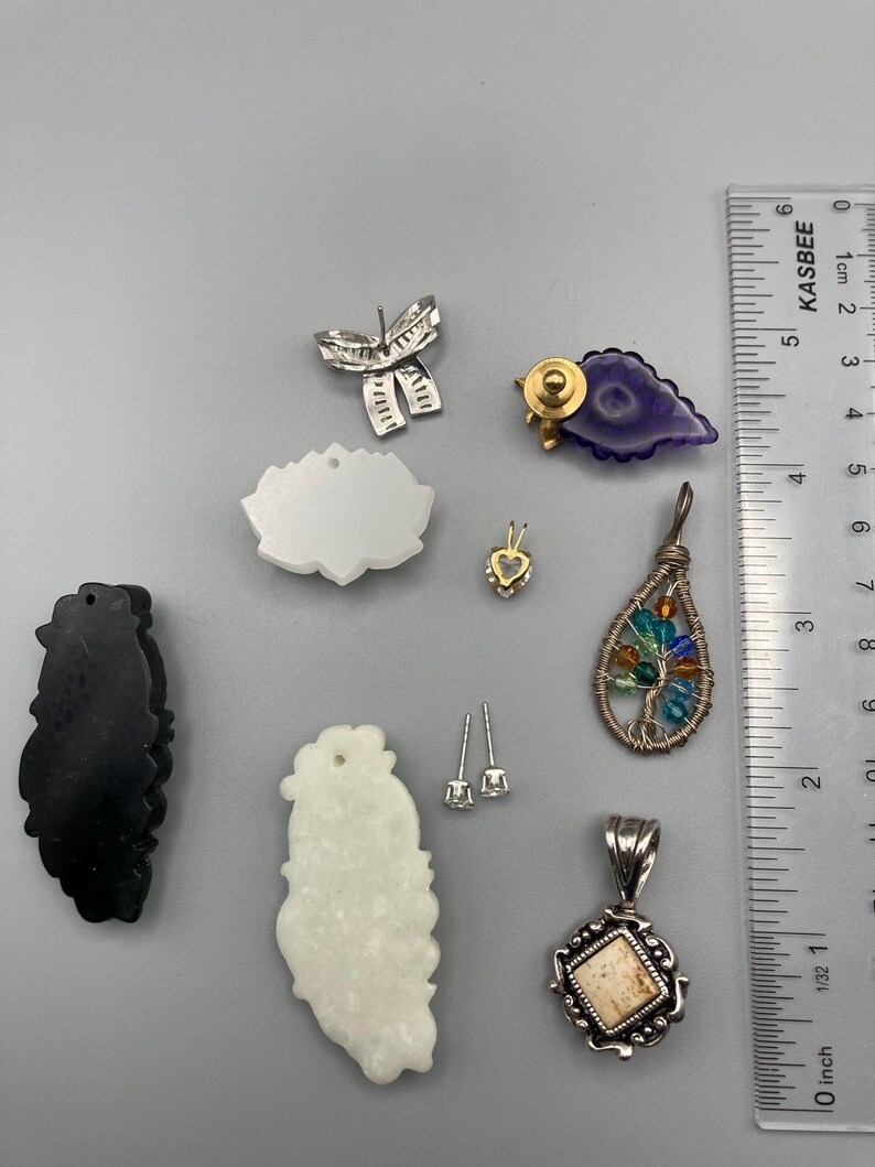 May include: A collection of jewelry pieces including a silver bow earring, a purple leaf-shaped pin, a white lotus-shaped pendant, a small heart pendant, a wire-wrapped tree pendant with colorful beads, a black and a white carved pendant, and stud earrings next to a ruler showing 3.5 inches.