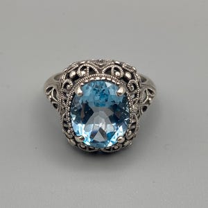 May include: A silver ring with an oval, light blue gemstone. The ring features an ornate, filigree design around the gemstone. The setting is detailed with small, round accents, creating a vintage aesthetic.