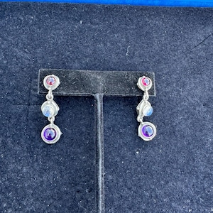 May include: A pair of silver dangle earrings with three gemstones. The top gemstone is red, the middle is white, and the bottom is purple.