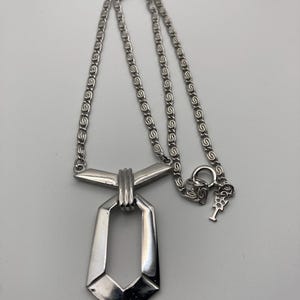 May include: A silver-toned necklace with a geometric pendant. The pendant has a rectangular shape with a pointed bottom and is connected to a chain with a decorative pattern. The necklace is displayed against a plain background.