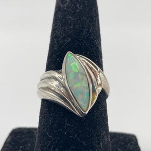 May include: A silver ring with an oval opal gemstone. The opal has a mix of green, white, and yellow colors. The ring's band has a wavy design on the sides, adding a unique touch to the jewelry.
