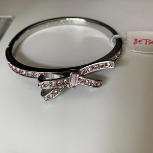 May include: A silver-tone hinged bangle bracelet featuring a central bow motif adorned with pink crystals. The bracelet is accented with a row of small pink crystals along the band and includes a tag labeled BETSEY JOHNSON. A ruler is visible.
