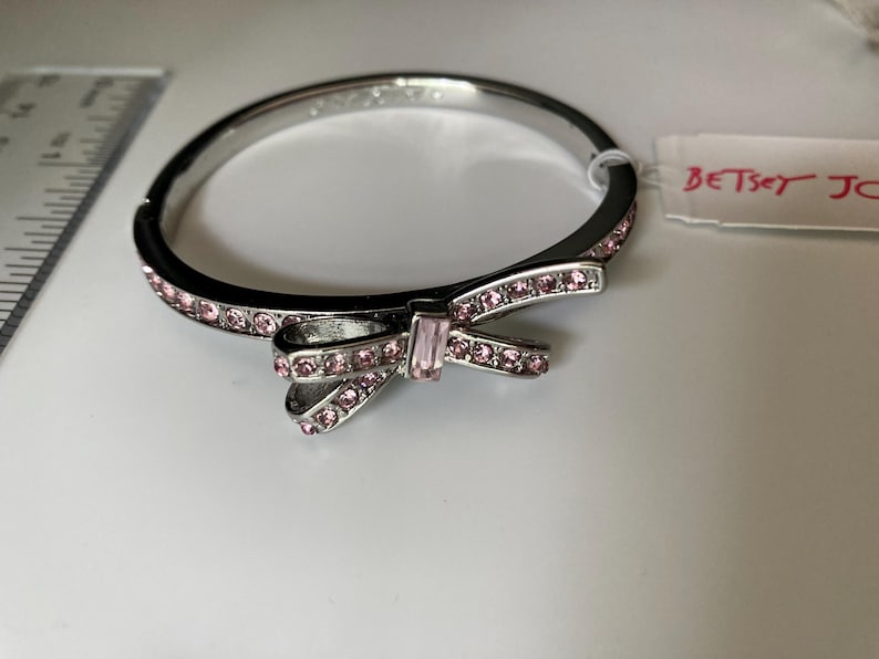 May include: A silver-tone hinged bangle bracelet featuring a central bow motif adorned with pink crystals. The bracelet is accented with a row of small pink crystals along the band and includes a tag labeled BETSEY JOHNSON. A ruler is visible.