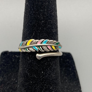 May include: A silver ring with a feather design, featuring yellow, pink, blue, and green enamel detailing. The ring wraps around the finger and has an adjustable design. The ring is a decorative piece.