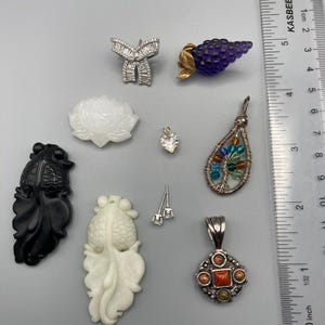 May include: A collection of jewelry pieces including a silver bow brooch, a purple grape-shaped pin, a white lotus pendant, a heart-shaped stone, a wire-wrapped tree pendant with colorful beads, a black fish carving, a white fish carving, and a silver pendant with a red stone, shown next to a ruler measuring up to 4 inches.