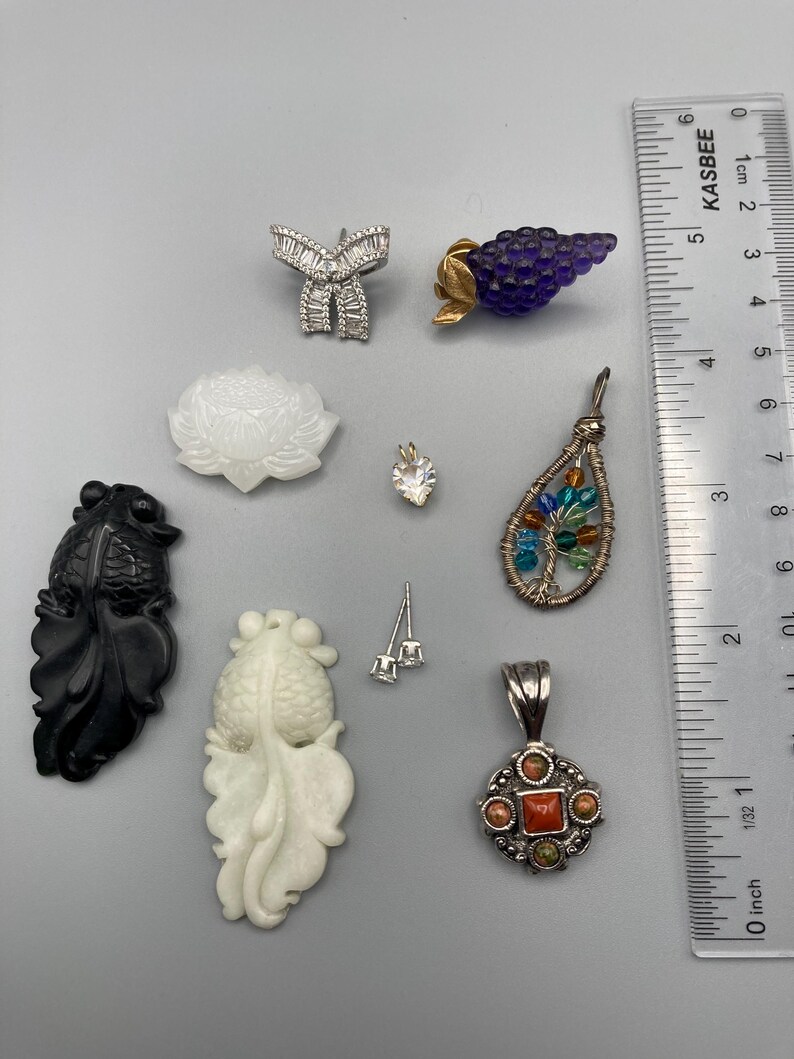 May include: A collection of jewelry pieces including a silver bow brooch, a purple grape-shaped pin, a white lotus pendant, a heart-shaped stone, a wire-wrapped tree pendant with colorful beads, a black fish carving, a white fish carving, and a silver pendant with a red stone, shown next to a ruler measuring up to 4 inches.
