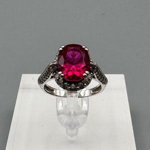 May include: A silver ring with a large oval-shaped red gemstone surrounded by smaller clear gemstones.