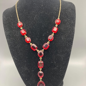 Op de afbeelding: Een goudkleurige ketting met een reeks facetgeslepen, robijnrode glazen stenen. De ketting heeft een Y-vormig ontwerp, met een hanger van afgestudeerde stenen die uitmondt in een grote druppelvormige edelsteen. Het wordt weergegeven op een zwarte mannequin.