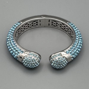 Può includere: Un braccialetto rigido in argento con un design di cristalli blu chiaro. Il braccialetto ha una chiusura a cerniera ed è decorato con piccoli cristalli rotondi.