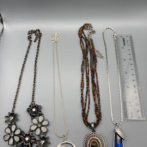 May include: Four necklaces displayed with a ruler measuring 6 inches in length. The first is a dark metal floral necklace with rhinestones. The second is a silver chain with a blue cabochon in a circle. The third has brown beads and a tiger eye pendant.