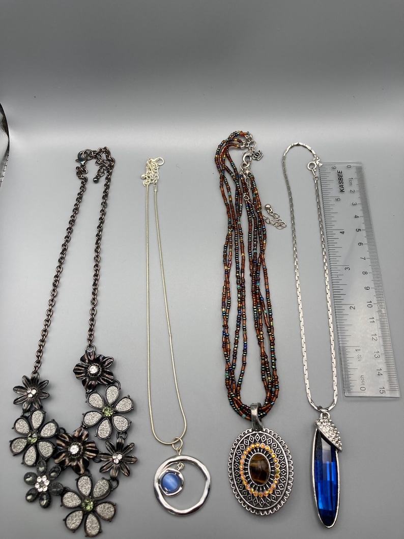 May include: Four necklaces displayed with a ruler measuring 6 inches in length. The first is a dark metal floral necklace with rhinestones. The second is a silver chain with a blue cabochon in a circle. The third has brown beads and a tiger eye pendant.