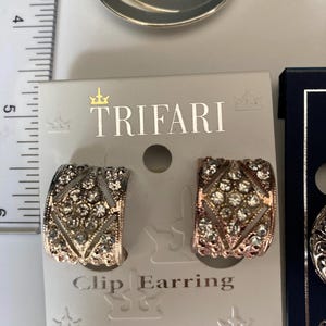 May include: A pair of Trifari clip earrings on a display card, featuring a curved, diamond-patterned design encrusted with clear rhinestones. A ruler is visible on the left, showing the earrings measure approximately 0.75 inches in height.