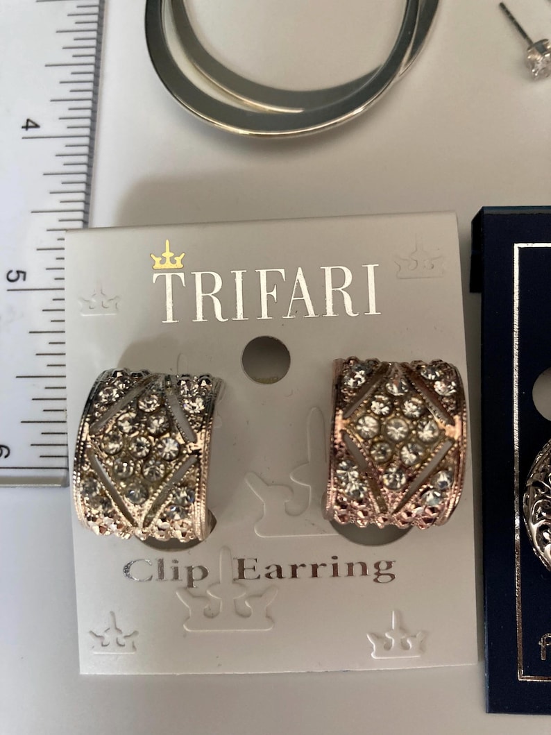 May include: A pair of Trifari clip earrings on a display card, featuring a curved, diamond-patterned design encrusted with clear rhinestones. A ruler is visible on the left, showing the earrings measure approximately 0.75 inches in height.