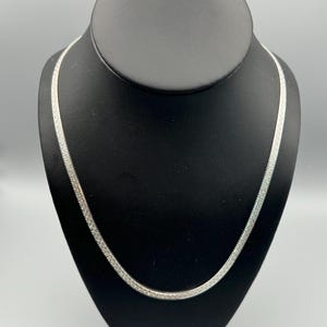 May include: A silver chain necklace on a black display stand. The chain is a simple, flat design.