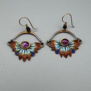 May include: A pair of artisan earrings with a fan-shaped design. The earrings feature a central purple gemstone, surrounded by a gold setting. The fan design has red, blue, and orange enamel details, with a silver-tone metal frame and gold-tone ear wires.