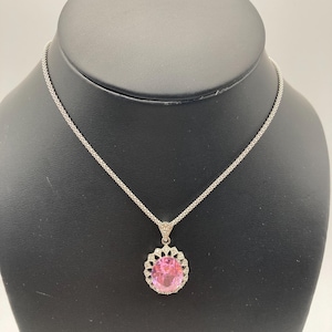 May include: A silver necklace with a large, oval pink gemstone pendant. The pendant is surrounded by a halo of smaller, clear stones and a decorative silver frame. The necklace is displayed on a black jewelry stand.
