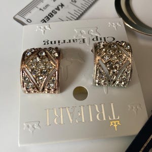 May include: A pair of Trifari clip earrings featuring a curved, wide band design encrusted with clear rhinestones in a geometric pattern. The earrings are attached to a white display card, with a ruler showing a width of approximately 0.75 inches.
