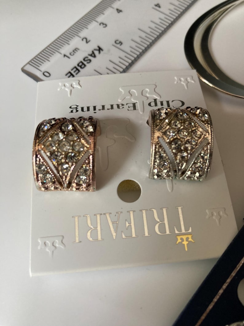 May include: A pair of Trifari clip earrings featuring a curved, wide band design encrusted with clear rhinestones in a geometric pattern. The earrings are attached to a white display card, with a ruler showing a width of approximately 0.75 inches.
