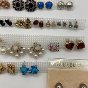 May include: A collection of various stud earrings displayed on white plastic grids. The earrings feature diverse designs, including floral, heart, and geometric shapes, with accents of blue, red, and pearl. Some earrings are in a clear plastic case with the brand name "RICHELIEU".
