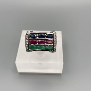 May include: A silver ring featuring three rows of gemstones. The top row has blue sapphires, the middle row has red rubies, and the bottom row has green emeralds. Small clear stones line the sides of the ring.
