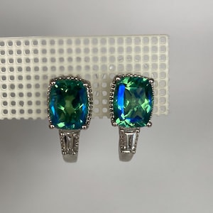 Puede incluir: Un par de pendientes plateados con grandes piedras preciosas cuadradas, iridiscentes verdes y azules. Las piedras preciosas están rodeadas por un borde de cuentas. Los pendientes tienen un diseño rectangular de corte baguette debajo de la piedra principal.
