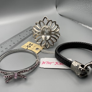 May include: A silver-tone ring with a clear oval center stone, a silver-tone hinged bangle with pink crystals and a bow detail, and a black braided bracelet with a silver-tone snake head clasp, shown with a ruler measuring up to 6 inches.
