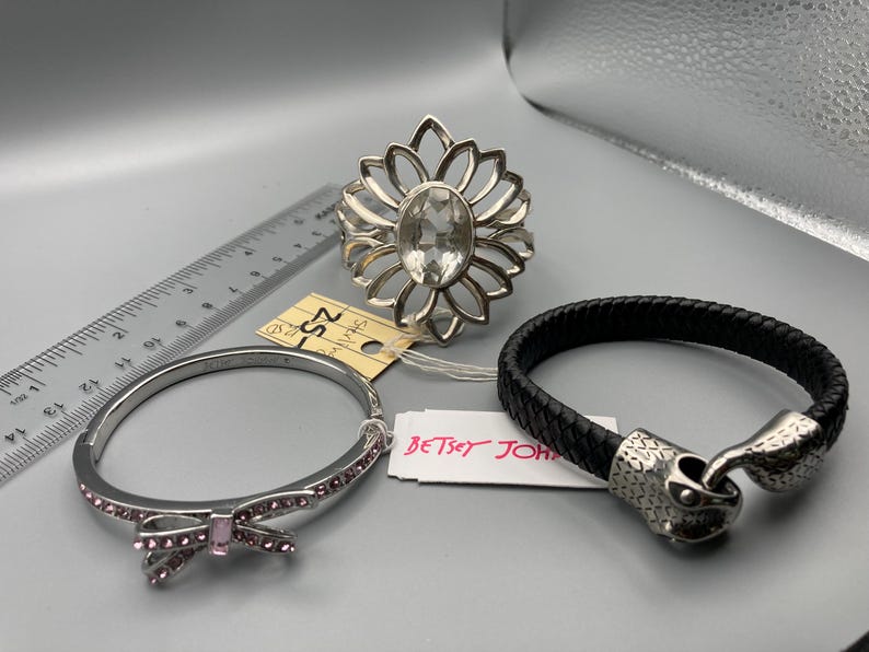 May include: A silver-tone ring with a clear oval center stone, a silver-tone hinged bangle with pink crystals and a bow detail, and a black braided bracelet with a silver-tone snake head clasp, shown with a ruler measuring up to 6 inches.