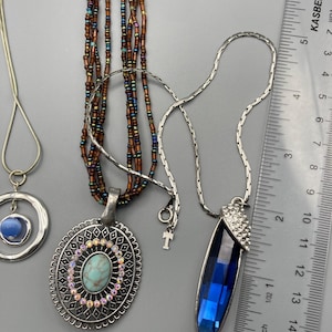 May include: Three necklaces are displayed next to a ruler. One features a blue stone in a silver circle, another has a multi-strand brown beaded chain with an oval turquoise-colored pendant, and the third has a long blue crystal pendant.