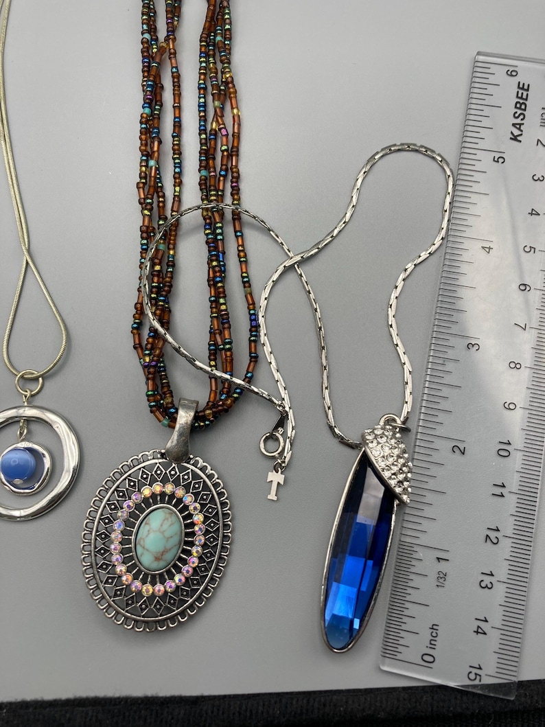 May include: Three necklaces are displayed next to a ruler. One features a blue stone in a silver circle, another has a multi-strand brown beaded chain with an oval turquoise-colored pendant, and the third has a long blue crystal pendant.