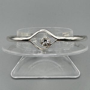 Puede incluir: Un brazalete plateado con una gema transparente en el centro. El brazalete presenta un diseño único con una abertura en forma de diamante alrededor de la gema. Se exhibe sobre un soporte transparente de varios niveles sobre un fondo gris.