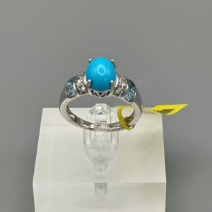 Puede incluir: Un anillo de plata con una gran piedra turquesa ovalada y brillante como pieza central. El anillo está adornado con piedras azules más pequeñas y gemas transparentes. El anillo se exhibe sobre un soporte transparente sobre un fondo gris.