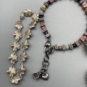 May include: A silver-toned bracelet featuring a chain of textured flower-shaped links and a second strand composed of multi-colored round beads interspersed with small, rectangular, etched silver-toned metal spacers and a heart-shaped clasp.