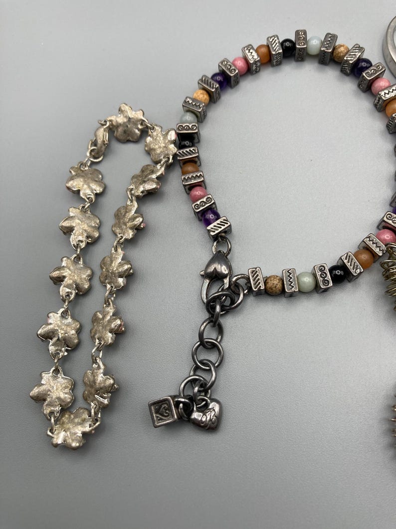 May include: A silver-toned bracelet featuring a chain of textured flower-shaped links and a second strand composed of multi-colored round beads interspersed with small, rectangular, etched silver-toned metal spacers and a heart-shaped clasp.