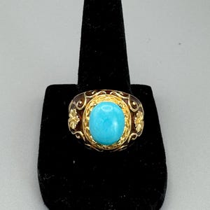 May include: A gold ring featuring a turquoise stone set in a bezel setting. The ring has an intricate design with floral motifs.