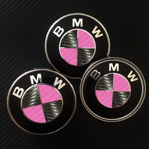 Op de afbeelding: Drie zwarte en zilveren BMW-autoemblemen met een roze en carbonfiber ontwerp.