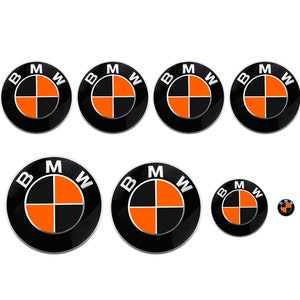 Puede incluir: Un conjunto de siete emblemas de coche BMW negros y naranjas de varios tamaños. Los emblemas son redondos y tienen un borde plateado.