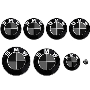 May include: A set of eight black and silver BMW car emblems of varying sizes. The emblems are round and feature the BMW logo with the letters "BMW" in silver and a black and white propeller design.
