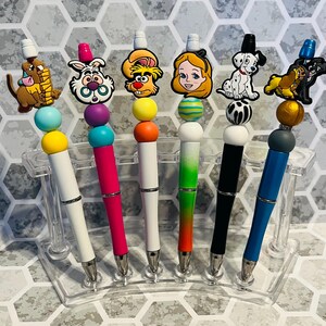 May include: Seven colorful pens with different cartoon character toppers. The pens are white, black, blue, green, pink, and orange. The toppers include a Dalmatian, a Disney princess, a cartoon dog, a cartoon cat, a cartoon rabbit, and a cartoon character with a crown.