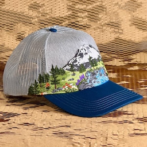 May include: A gray trucker hat with a blue brim and a mountain scene embroidered on the front. The mountain scene includes trees, flowers, and a waterfall. The hat has a mesh back.